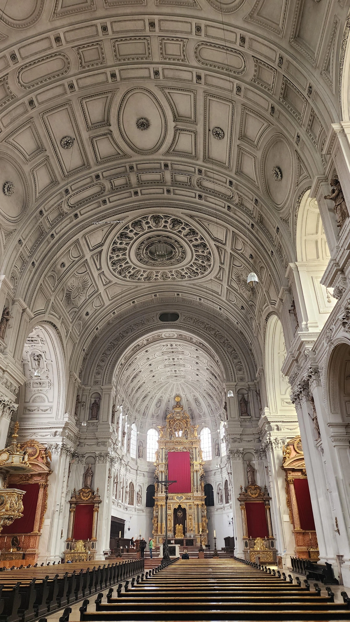 Looking down the nave with the gigantic barrel vault of the ceiling overhead, its only decoration being raised edges or frames dividing it into oval and rectangular sections. The ceiling and the rectangular arched columns supporting it are white. To the left and right are large red and gold altarpieces of side chapels. At center in the distance is the very large church altarpiece, with gold ornament and red cloth background.